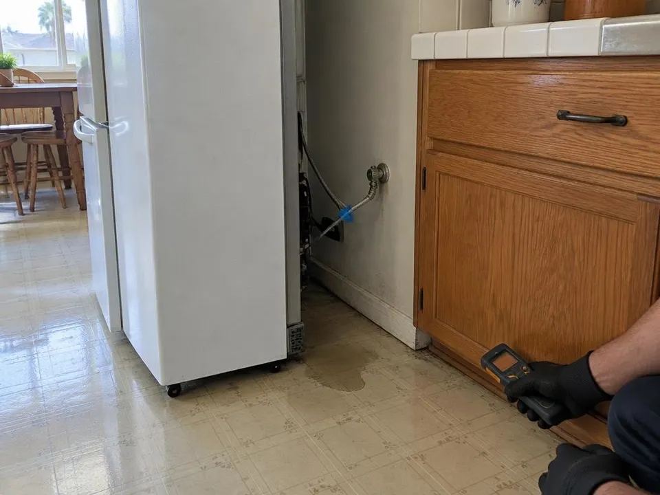 Generated illustration of a gloved hand holding a moisture meter near a cabinet base beside a refrigerator supply line and a small puddle